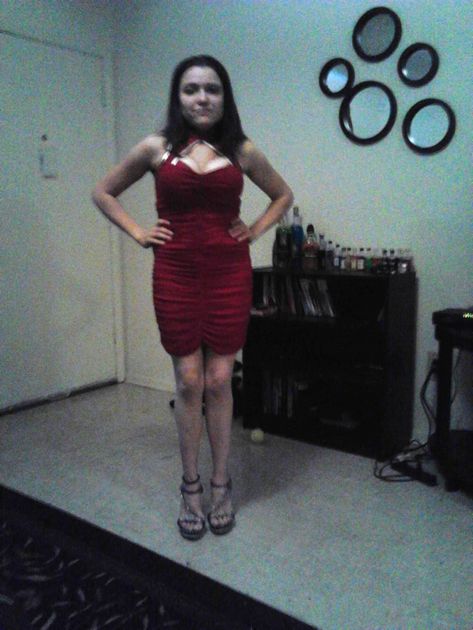 Woman in glasses smiles, wearing blue sweater over dark teal dress, arms at sides, in a room.