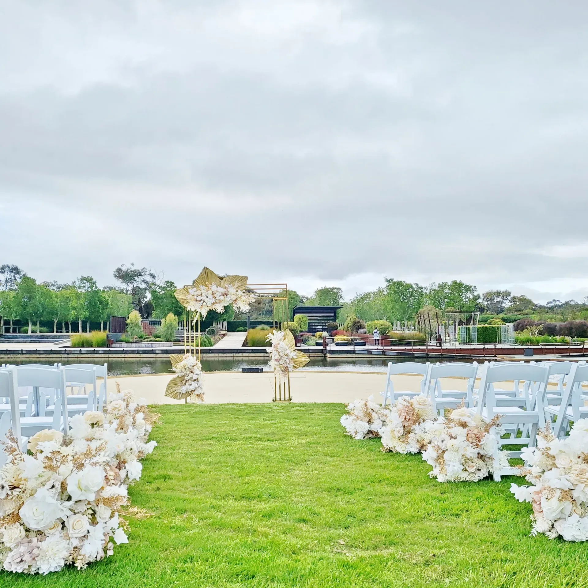Outdoor wedding aisle with white chairs and floral arrangements facing a lake and garden backdrop