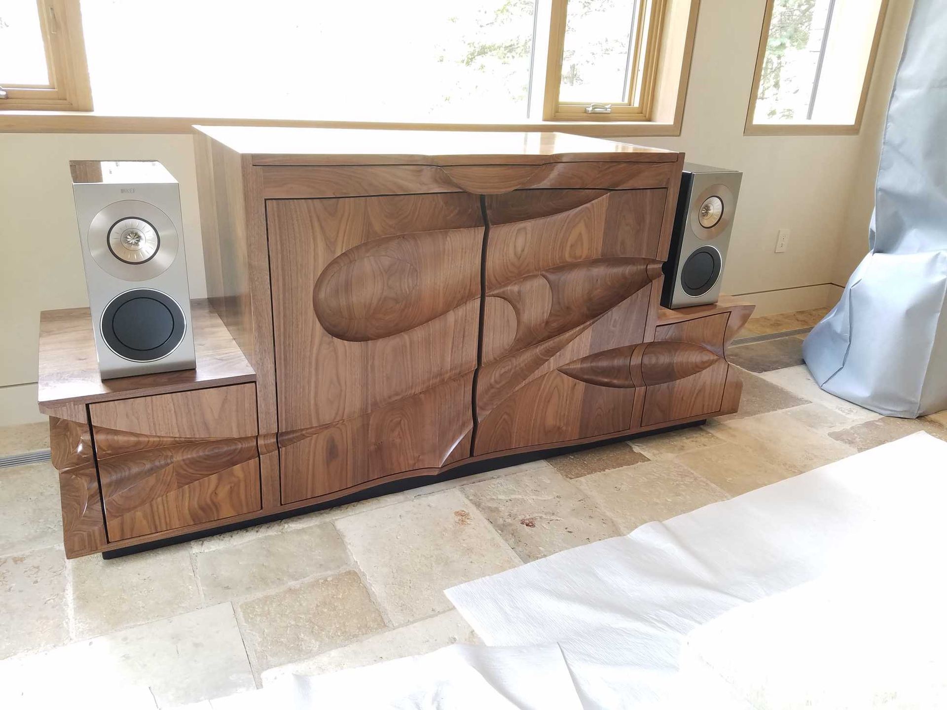 Wooden cabinet with carved design, speakers on each side, in a room with a window.