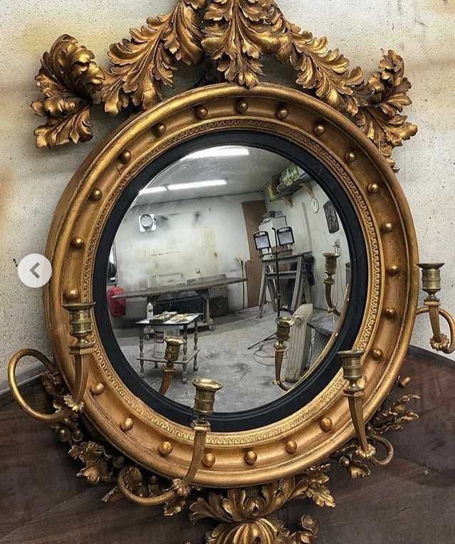 Gold-framed circular mirror with candle holders, ornate detailing, reflects a cluttered workshop space.