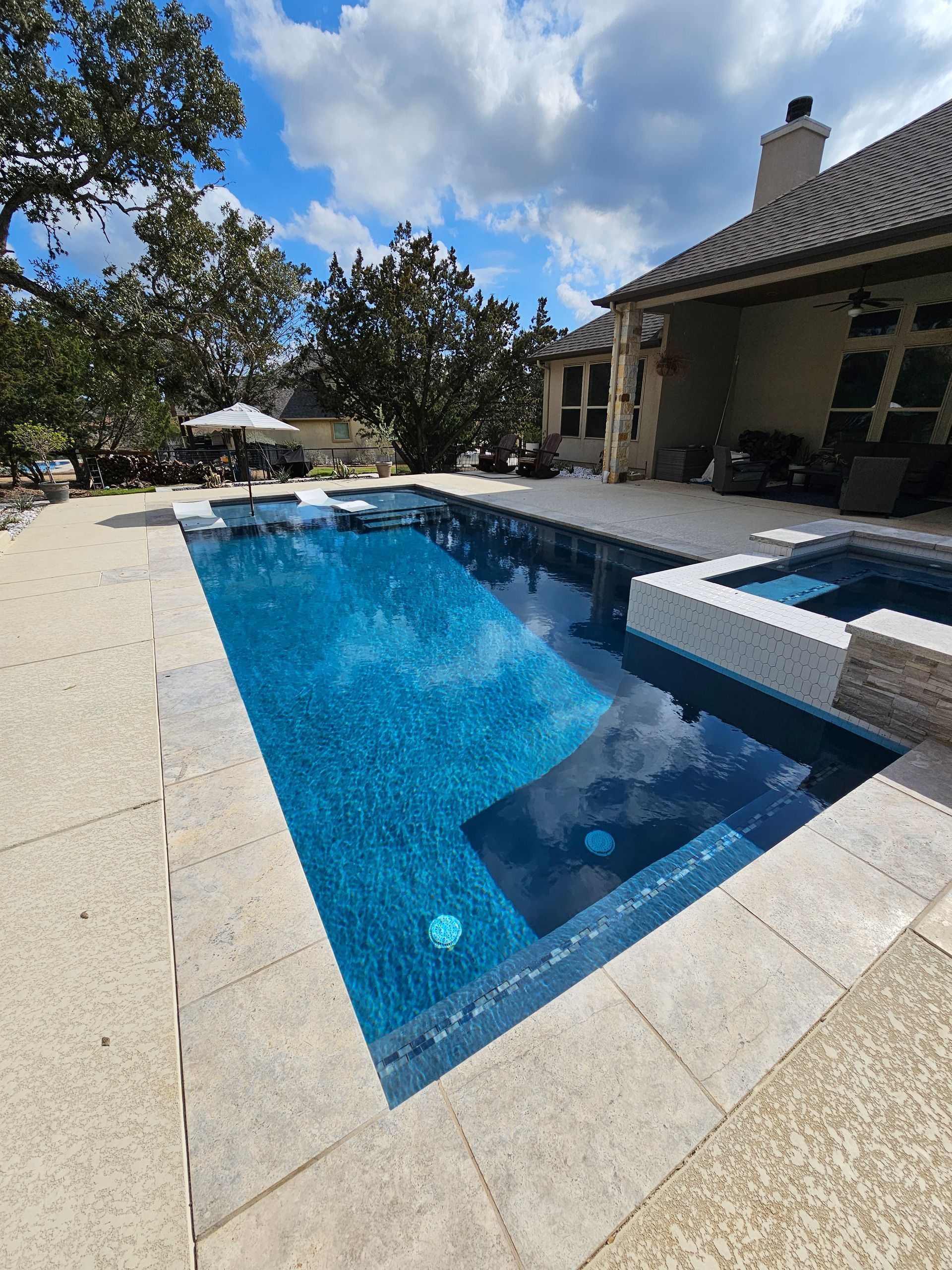Pool with blue tiled interior, connected spa, and light-colored stone patio, under a cloudy sky.