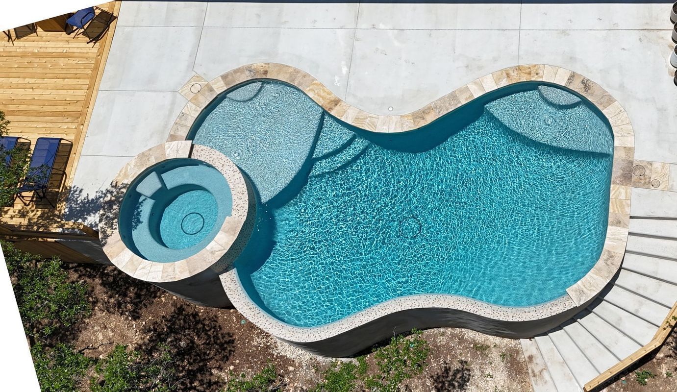 Pool with blue tiled interior, connected spa, and light-colored stone patio, under a cloudy sky.
