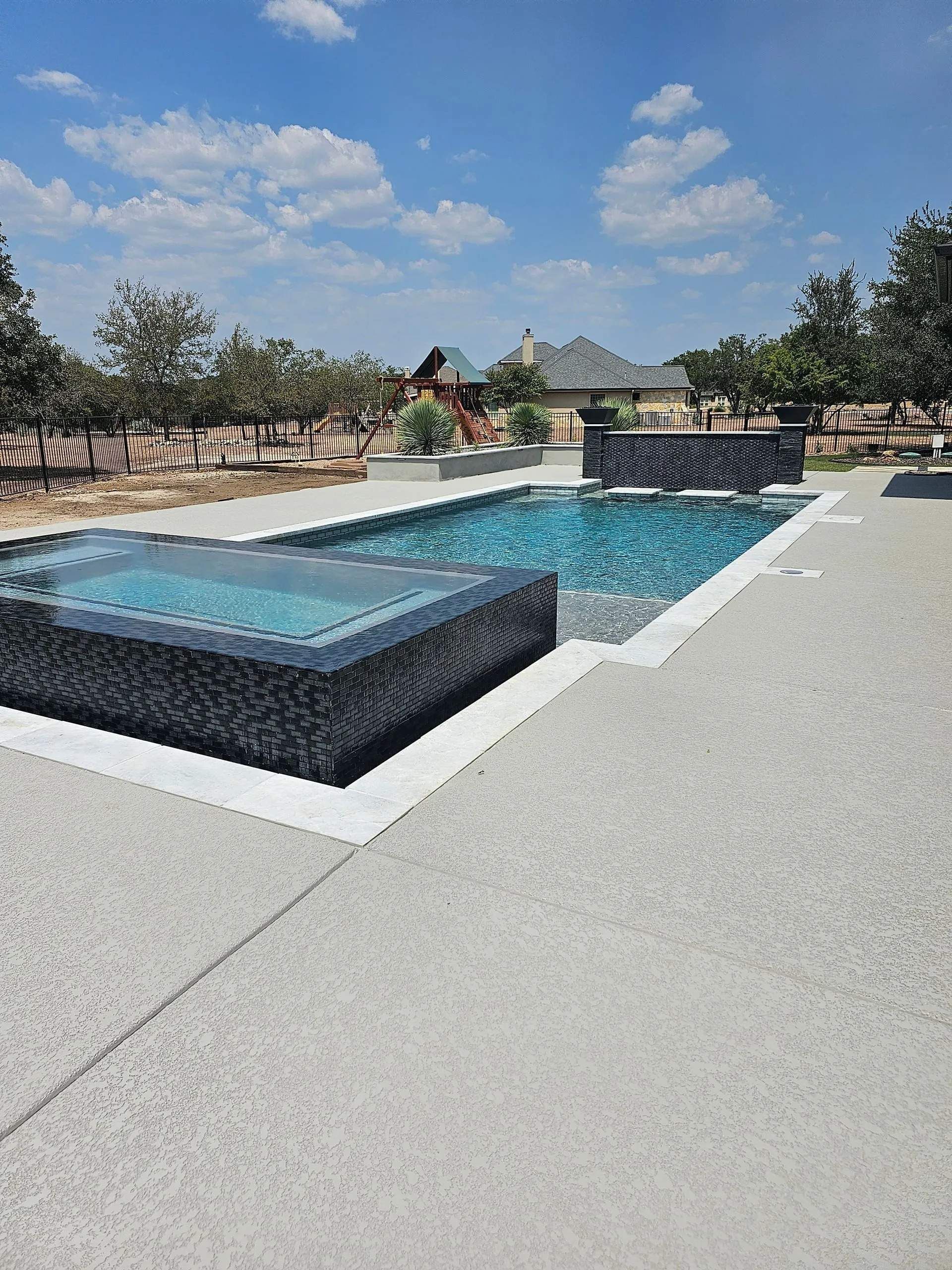Pool with blue tiled interior, connected spa, and light-colored stone patio, under a cloudy sky.