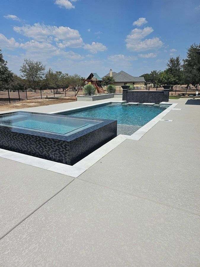 Pool with blue tiled interior, connected spa, and light-colored stone patio, under a cloudy sky.