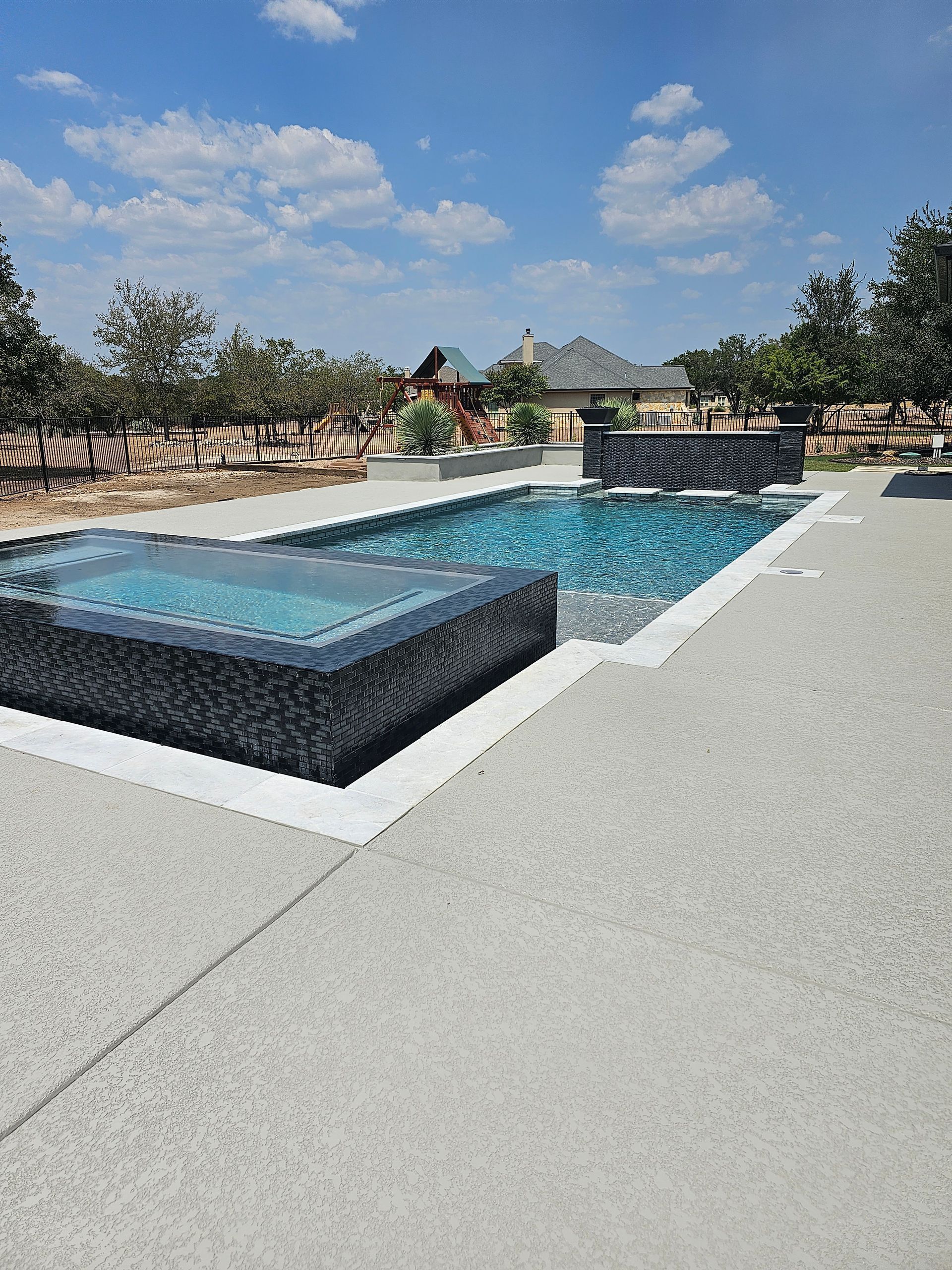 Pool with blue tiled interior, connected spa, and light-colored stone patio, under a cloudy sky.