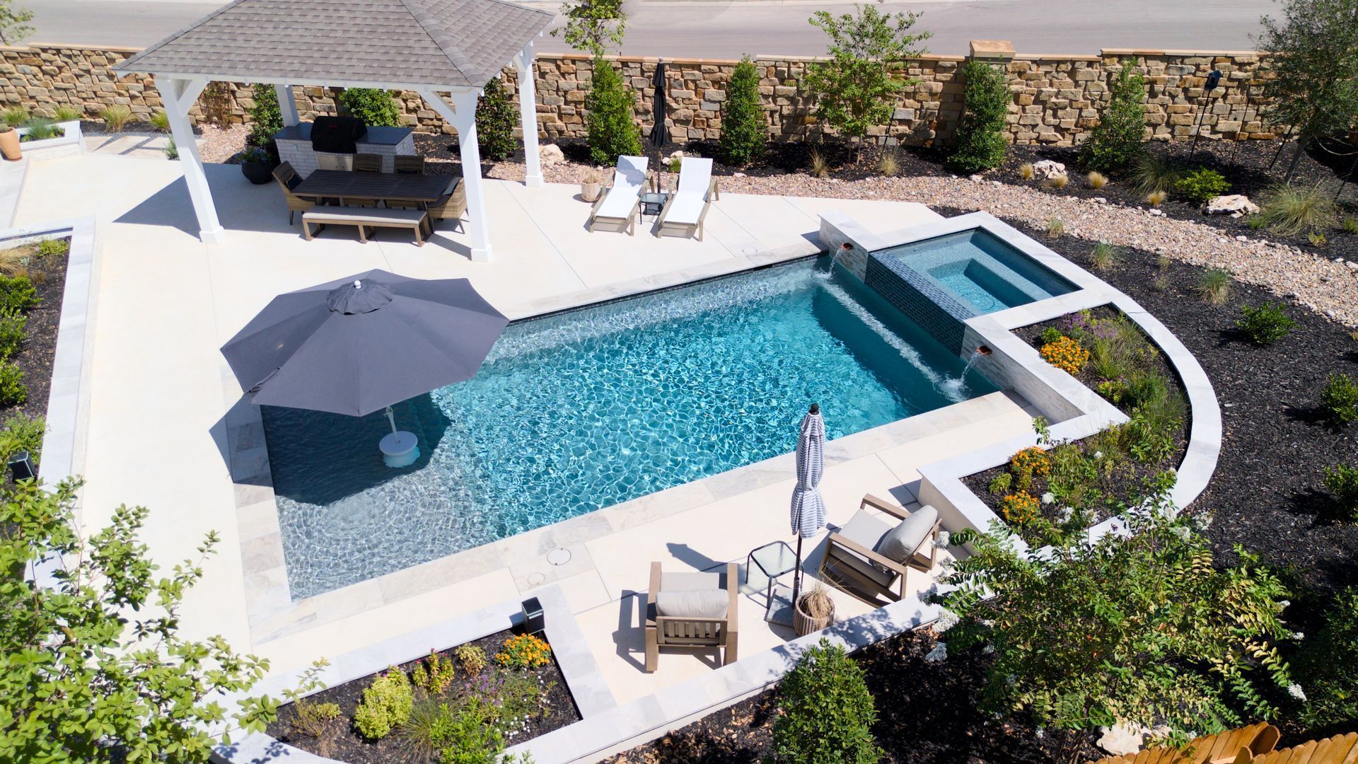 Pool with blue tiled interior, connected spa, and light-colored stone patio, under a cloudy sky.