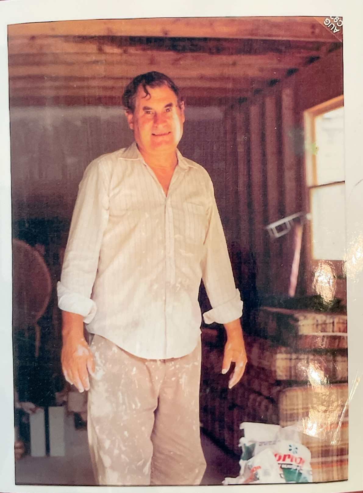 Man in light-colored shirt and pants, covered in white dust, smiles in a room with exposed wooden beams.