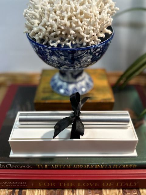 A silver pen with a black ribbon in a white box, stacked on books, topped with coral in a blue bowl.