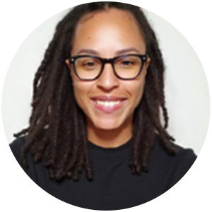 Person with locs and glasses smiles, in a black shirt, in a white-walled room.