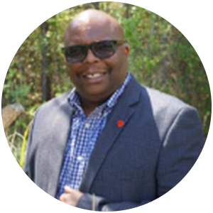 Man wearing sunglasses, blazer, and checkered shirt, smiling outdoors in front of foliage.