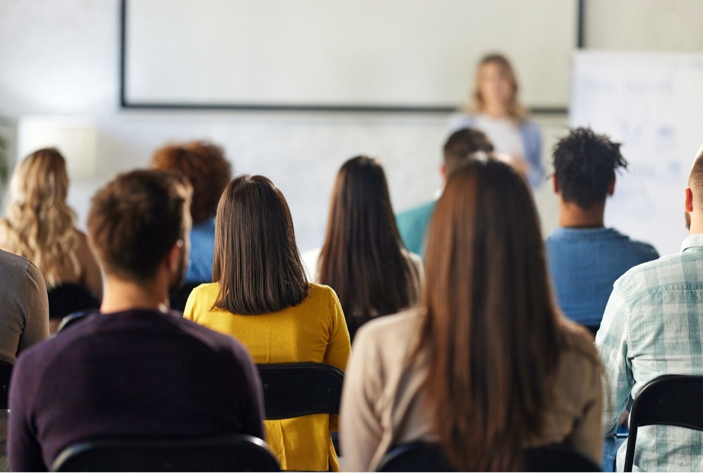 Audience seated, facing a woman presenting in a classroom.
