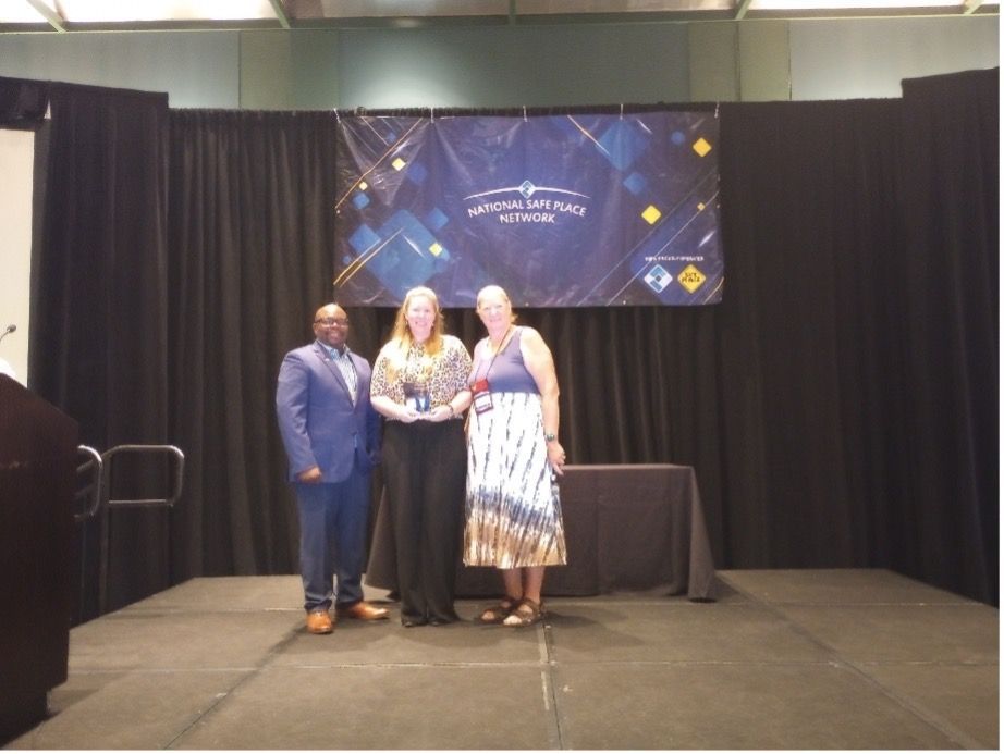 Three people on a stage with a banner, holding an award. Person in the middle is holding the award.