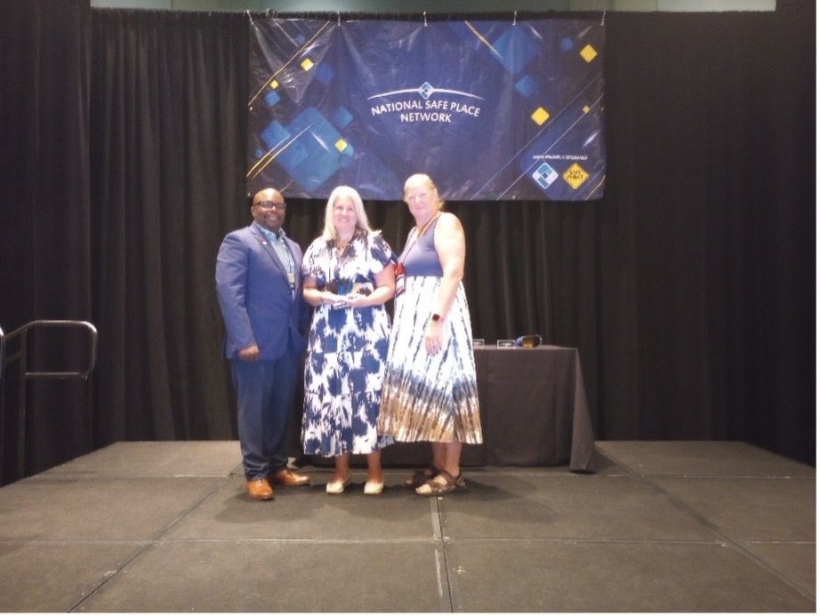 Three people on a stage in front of a banner. A man in a suit, two women in dresses, and a table.