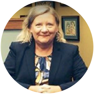 Woman in a blazer, smiling, sitting in a room with a framed picture on the wall.