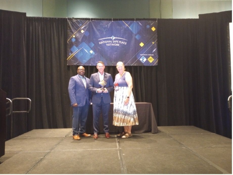 Three people on a stage: a man holds a trophy, flanked by a man in a blue suit and a woman in a tie-dye dress.