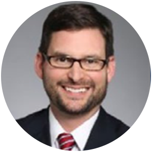 Man with glasses, smiling in a suit and tie.  Background is a soft gray.