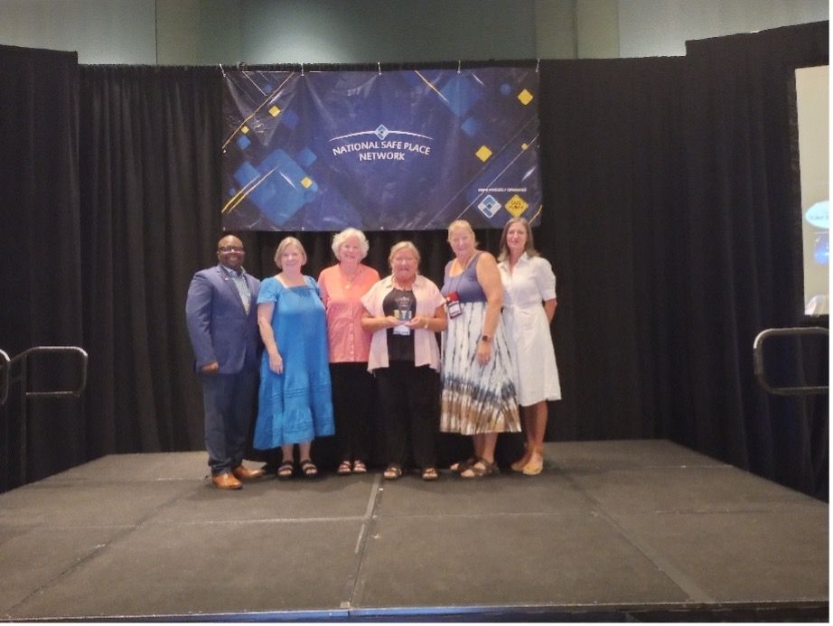 People on stage, holding an award. Backdrop with event logo. Dark stage, attendees are smiling.