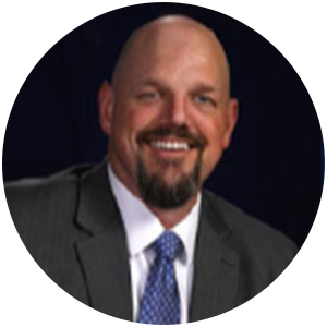Bald man in a suit smiles, close-up shot against a dark blue background.