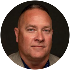 Man in a blazer and blue shirt smiles slightly, against a dark background.