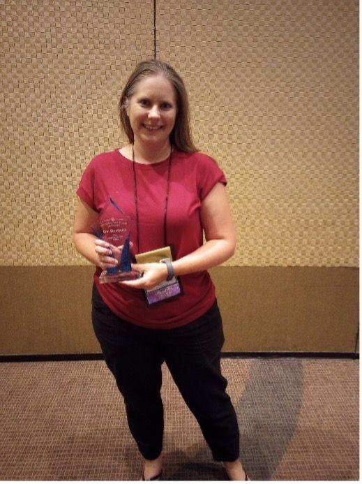 Woman in red shirt and black pants holding a star-shaped award, standing in front of a textured wall.