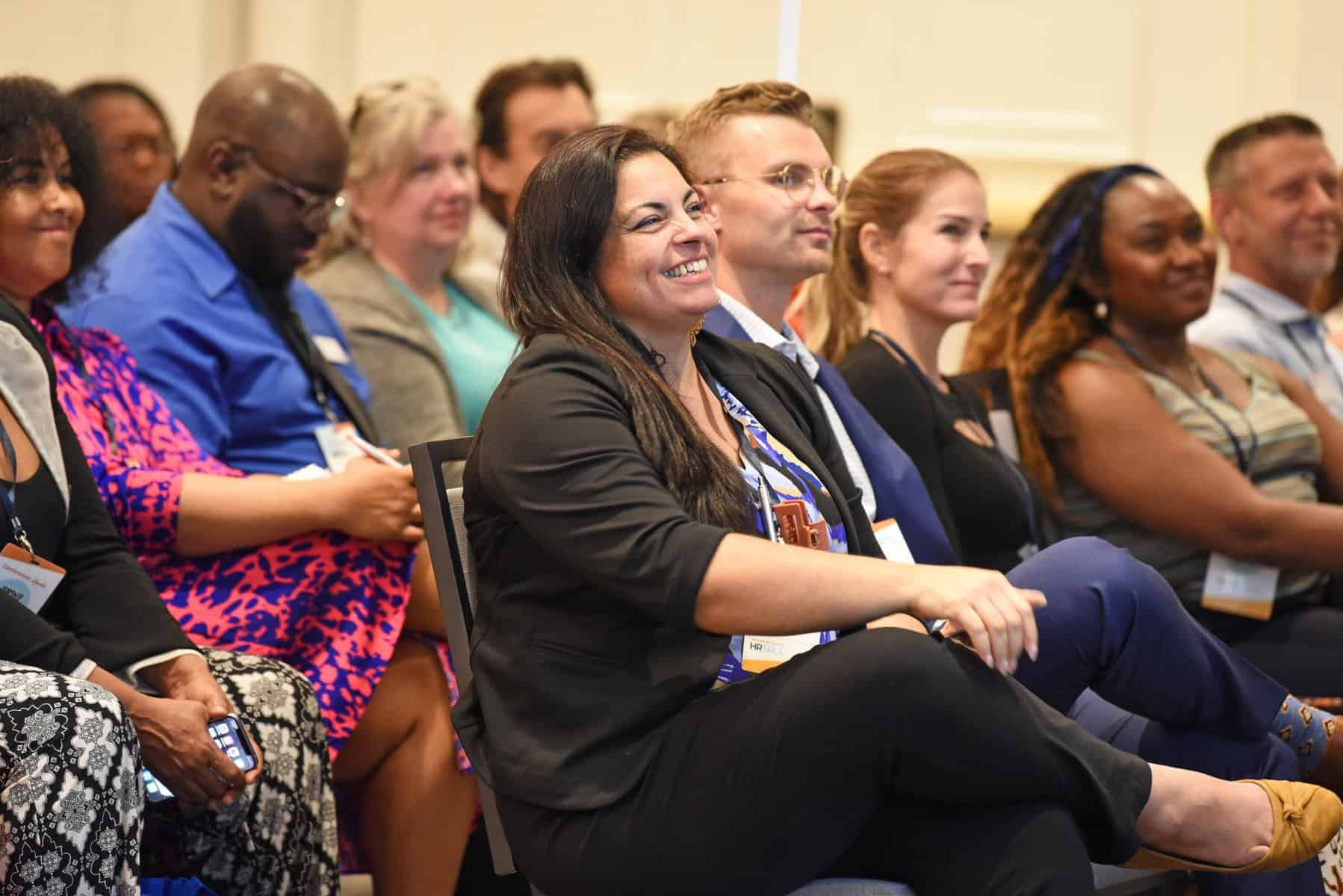 Audience seated, smiling and attentive at an event. People in various business attire.