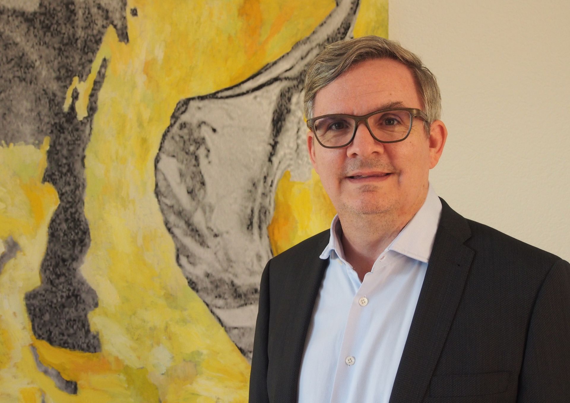 Ein Mann mit Brille, Blazer und offenem Hemdkragen lächelt; er steht neben einem gelb-grauen abstrakten Gemälde an einer weissen Wand.