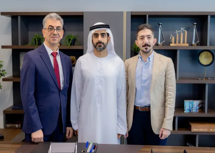 Three men in an office: one in a white thobe, one in a blue suit, and one in a tan jacket, posing for a photo.