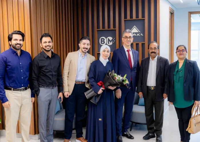 Group of people in an office setting; woman in blue dress holds flowers; smiling faces.