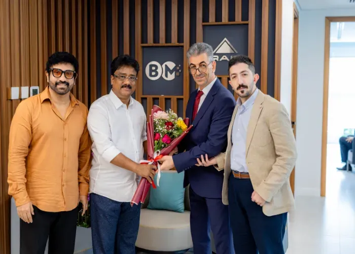 Four men in an office exchanging a flower bouquet. Two are smiling, in front of a logo on a wall.