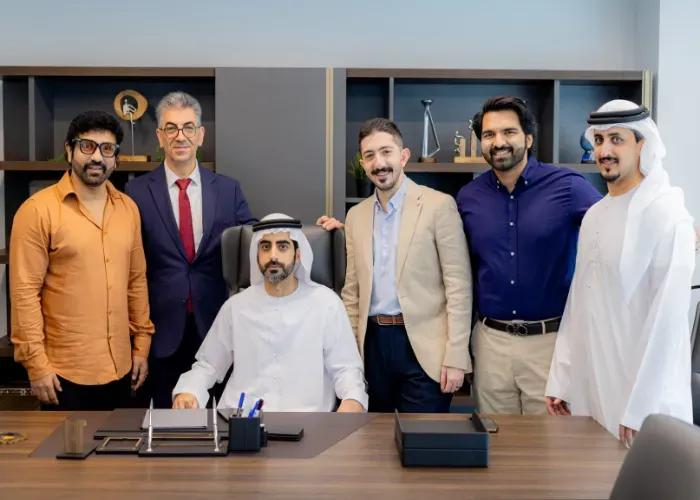 A group of six men in a modern office setting. Some are wearing traditional Arab attire, and others are in business casual.