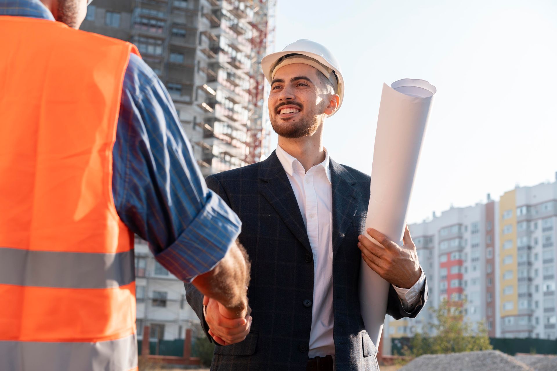 Construction worker and architect shaking hands, smiling. Building site in background.