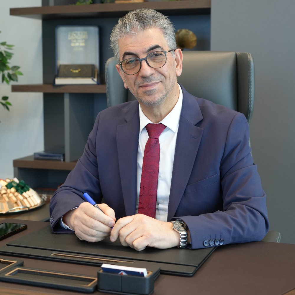 Man in suit, seated at desk, holding pen, looking at camera. Office setting with bookshelf.