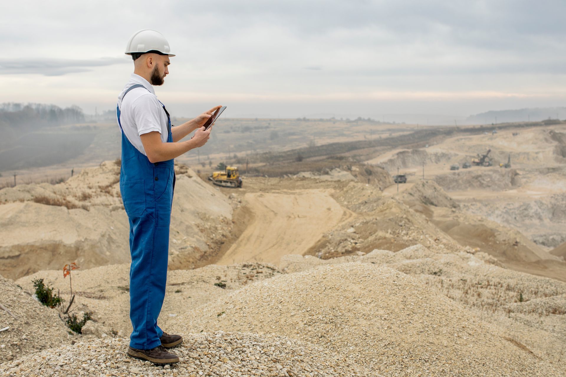 Construction worker in blue overalls and white hard hat using a tablet, overlooking a quarry.