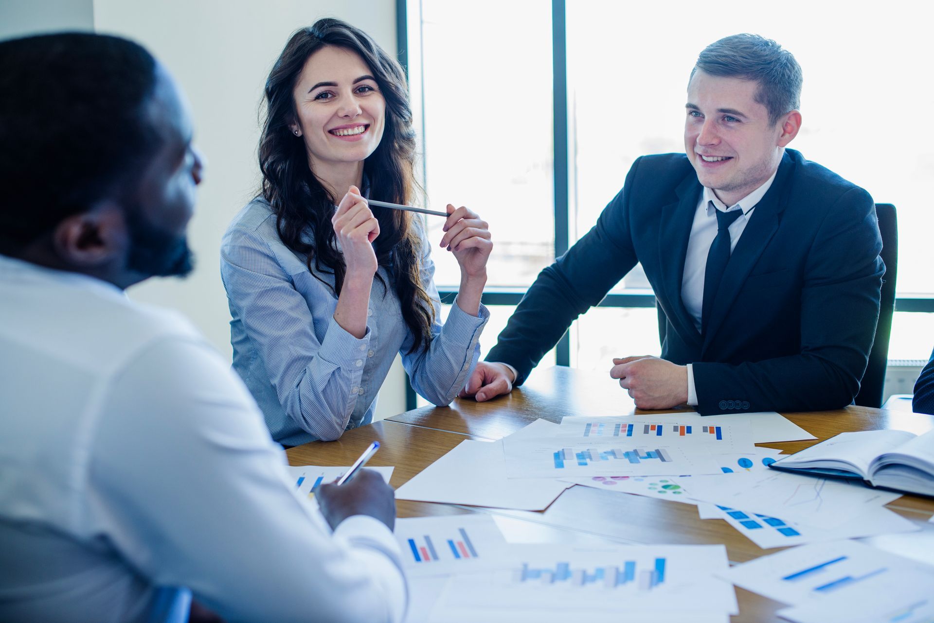 Three business colleagues at a table discussing charts. Smiling woman and man in suits.
