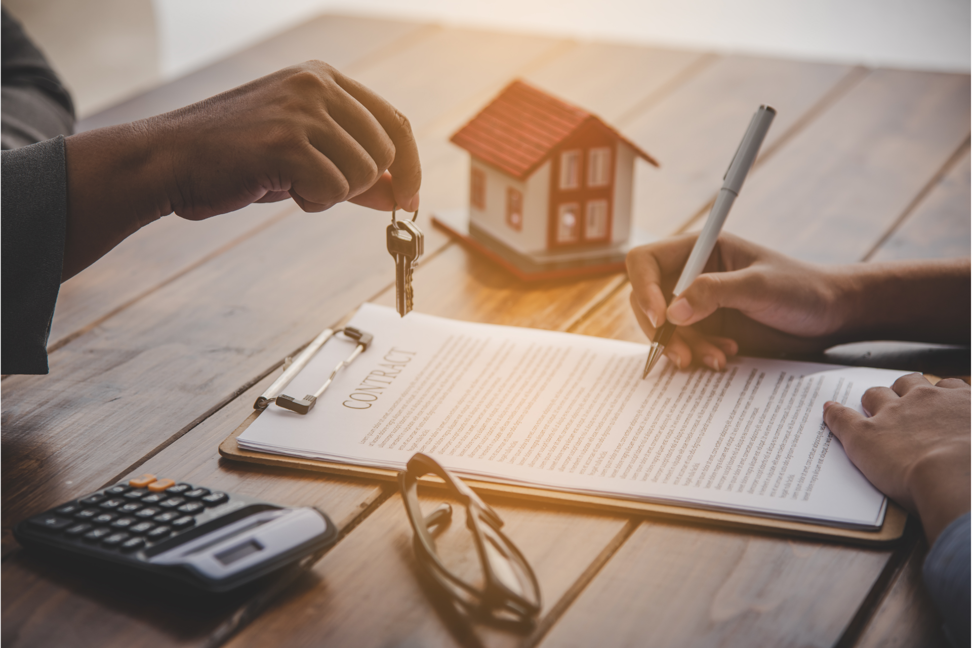 Hands exchanging house keys over a clipboard with a contract, calculator, and model house on a desk