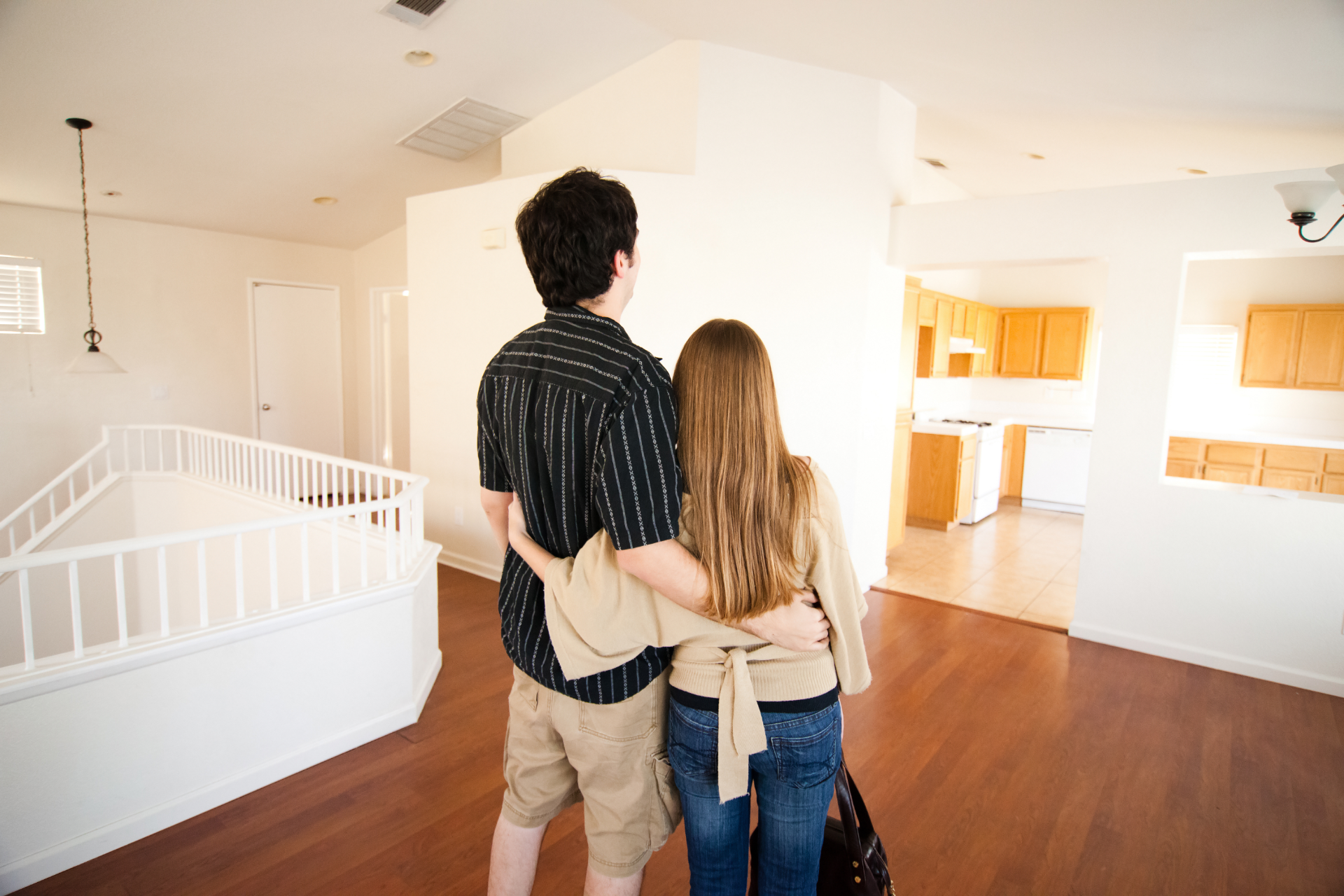 Couple standing in a bright empty room, looking into a home interior with wood floors and white walls.