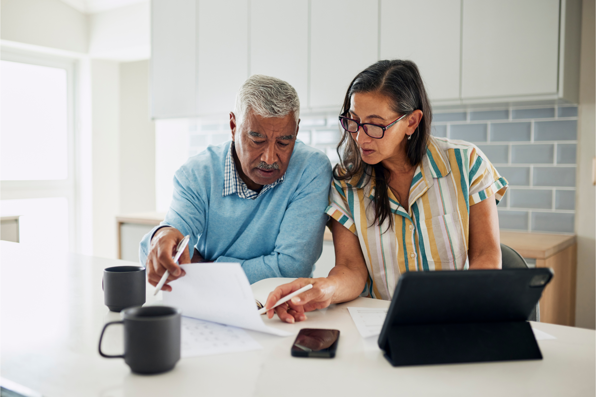 Two people reviewing papers at a kitchen table with a laptop and coffee mugs