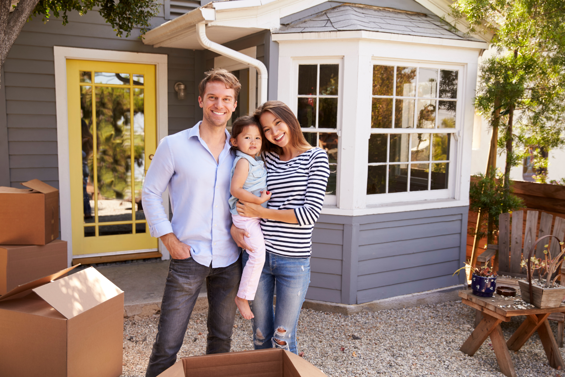 Family standing outside a small house with a yellow door and bay window, beside moving boxes