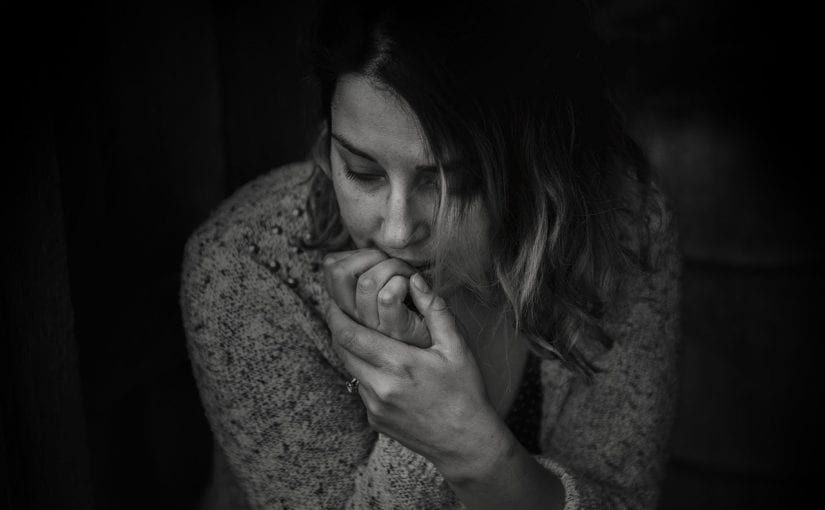 Woman in black and white, with her hands clasped in front of her face, looking down; appears distressed.