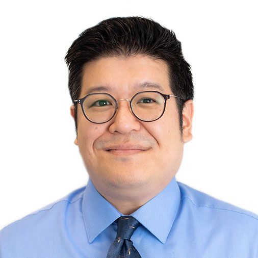 A man wearing glasses and a blue shirt and tie is smiling.