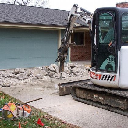 A bobcat excavator is breaking concrete in front of a house.