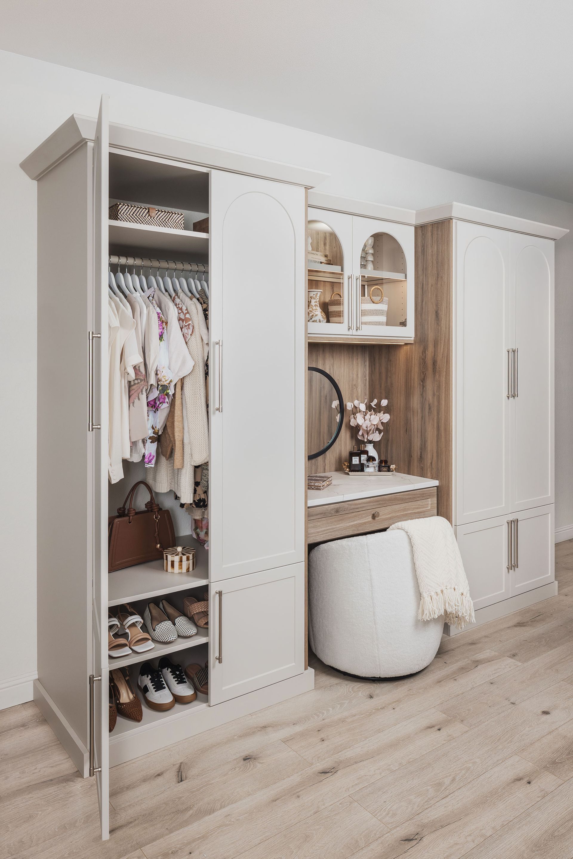 White walk-in closet with hanging clothes, shelves, mirrored vanity, and a small ottoman on wood flooring.