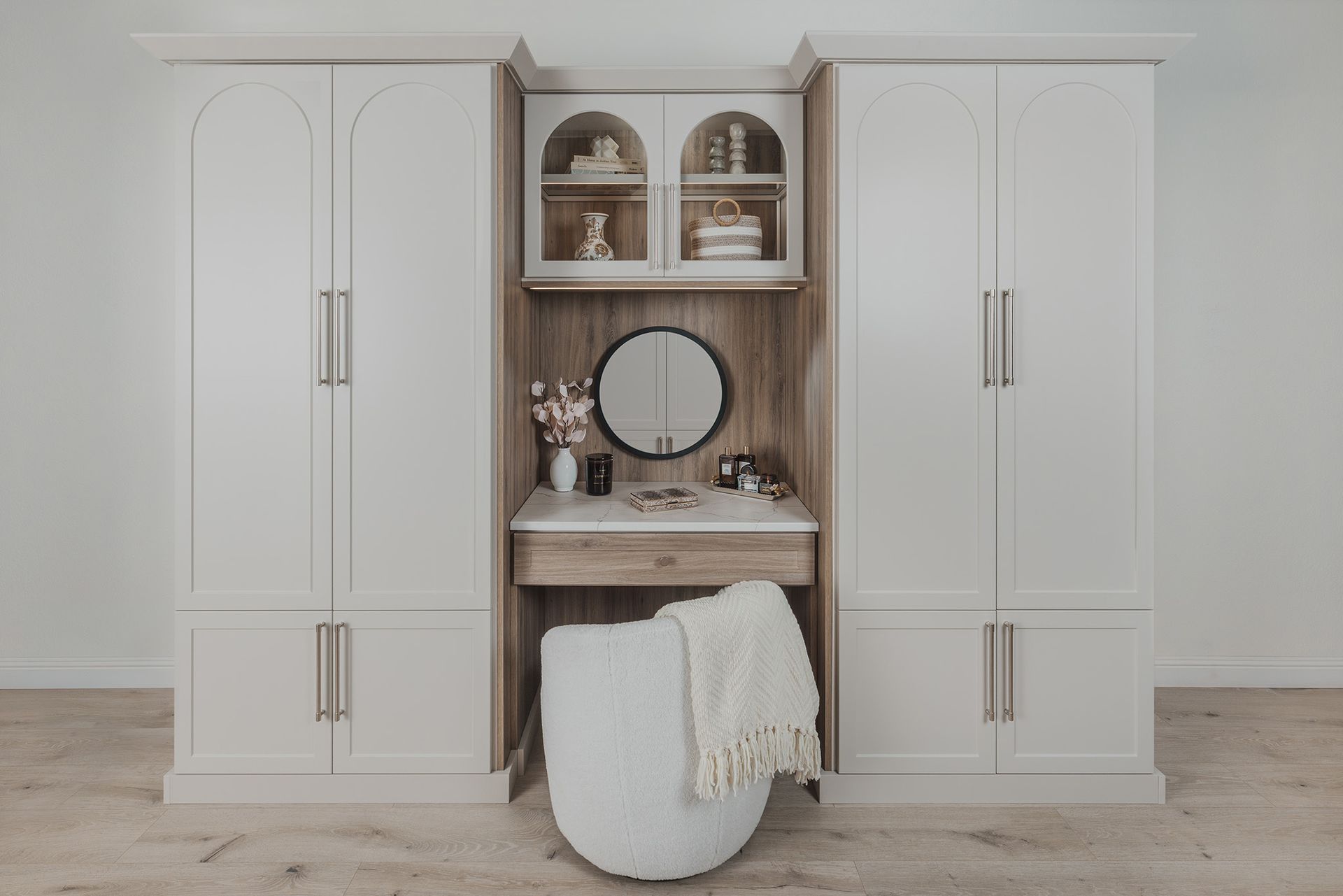 Built-in white wardrobe and vanity with round mirror, decor shelves, and a white ottoman in a neutral room.