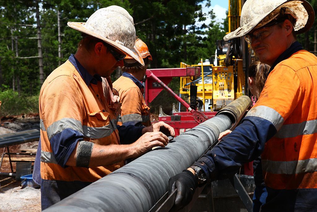 Workers in orange coveralls examine a long core sample outdoors near drilling equipment.