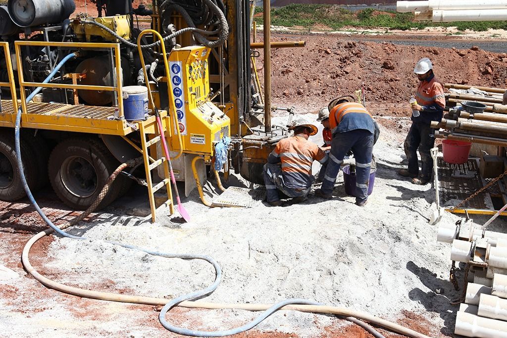 Workers operating drilling equipment at a construction site. Yellow machinery, reddish earth, and bright sky.