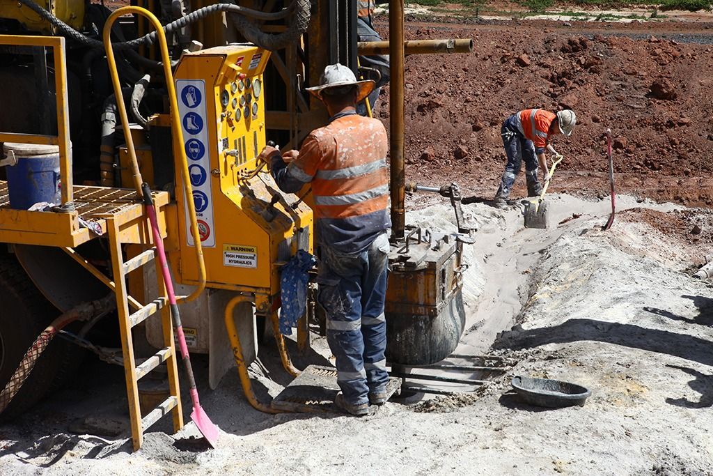 Two construction workers at a drilling site. One operates a machine, the other shovels. Outdoors in sunlight.