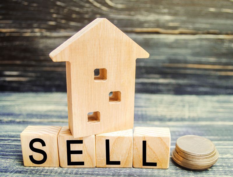 A wooden house is sitting on top of wooden blocks with the word sell written on them.