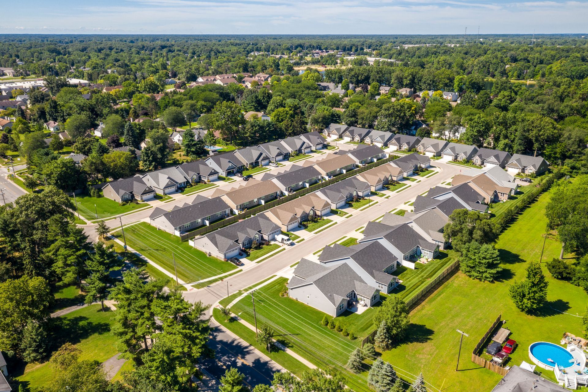 Aerial view of a suburban neighborhood with rows of houses, green lawns, and trees under a sunny sky.