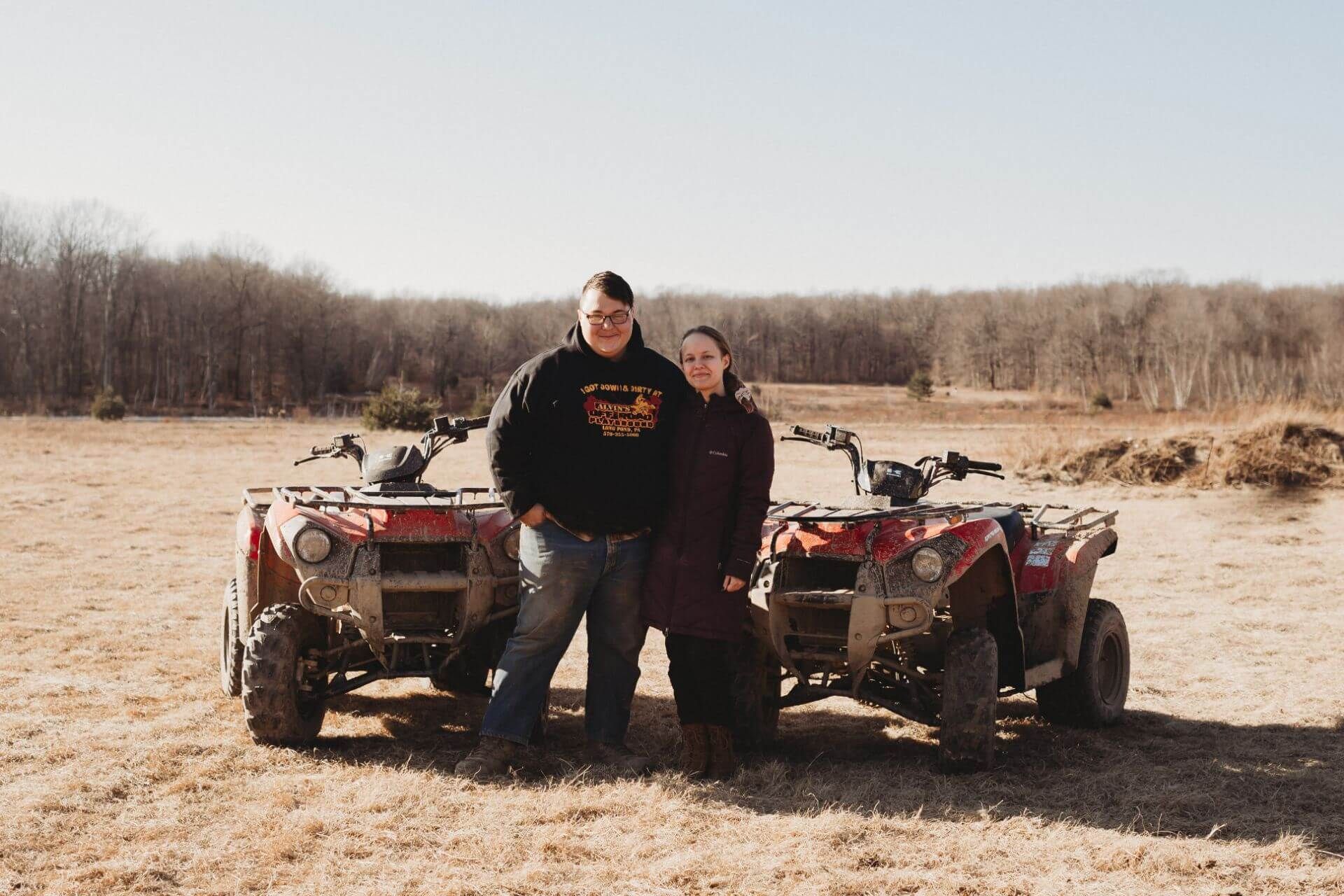 A man and a woman are standing next to two atvs in a field.