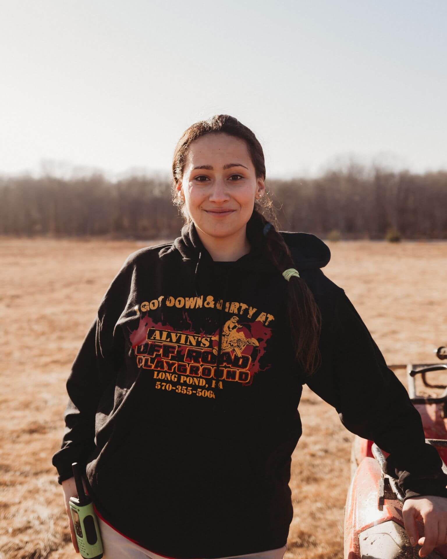 A woman in a black hoodie is standing in a field holding a bottle of beer.